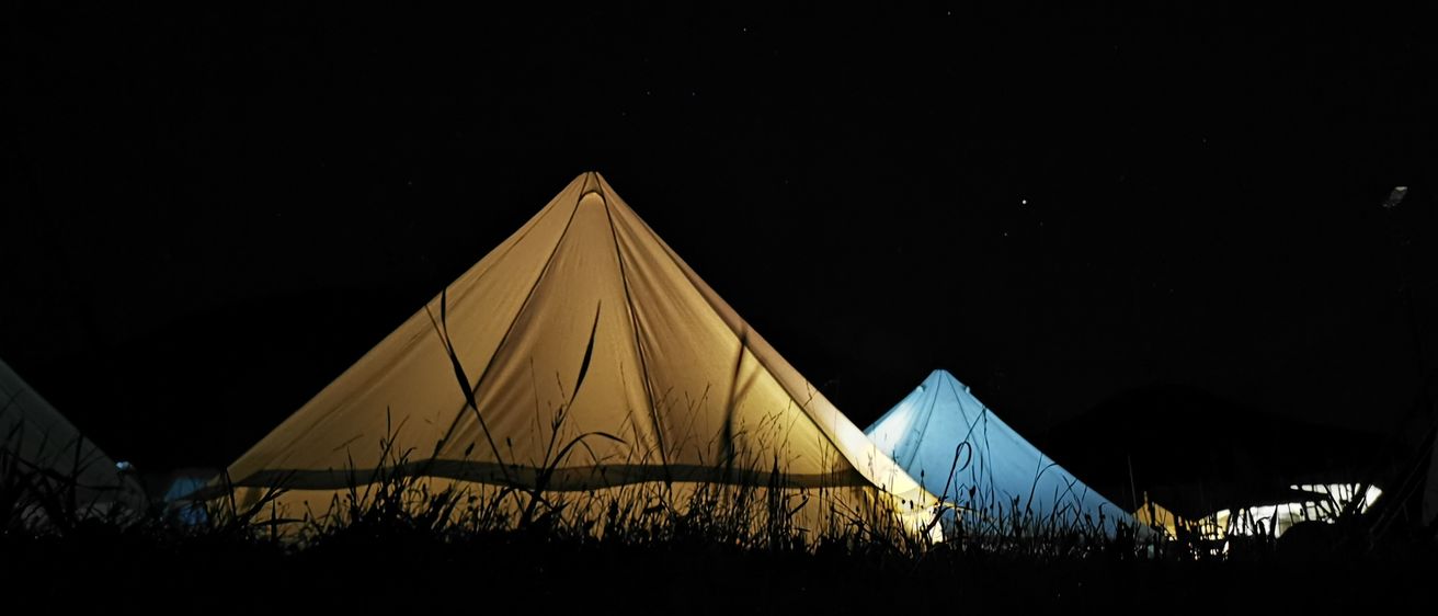 Zwei Zelte sind in einem Grasfeld bei Nacht aufgestellt und von einer Lichtquelle beleuchtet. Die Zelte sind beige und blau und werfen Schatten auf den Boden.