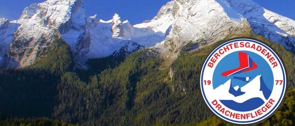 Eine verschneite Bergkette unter einem klaren blauen Himmel. Im Vordergrund befindet sich ein Logo mit 'BERCHTESGADEN' und '19'. Das Logo zeigt eine Person in einem roten Hut und einem Fallschirm.