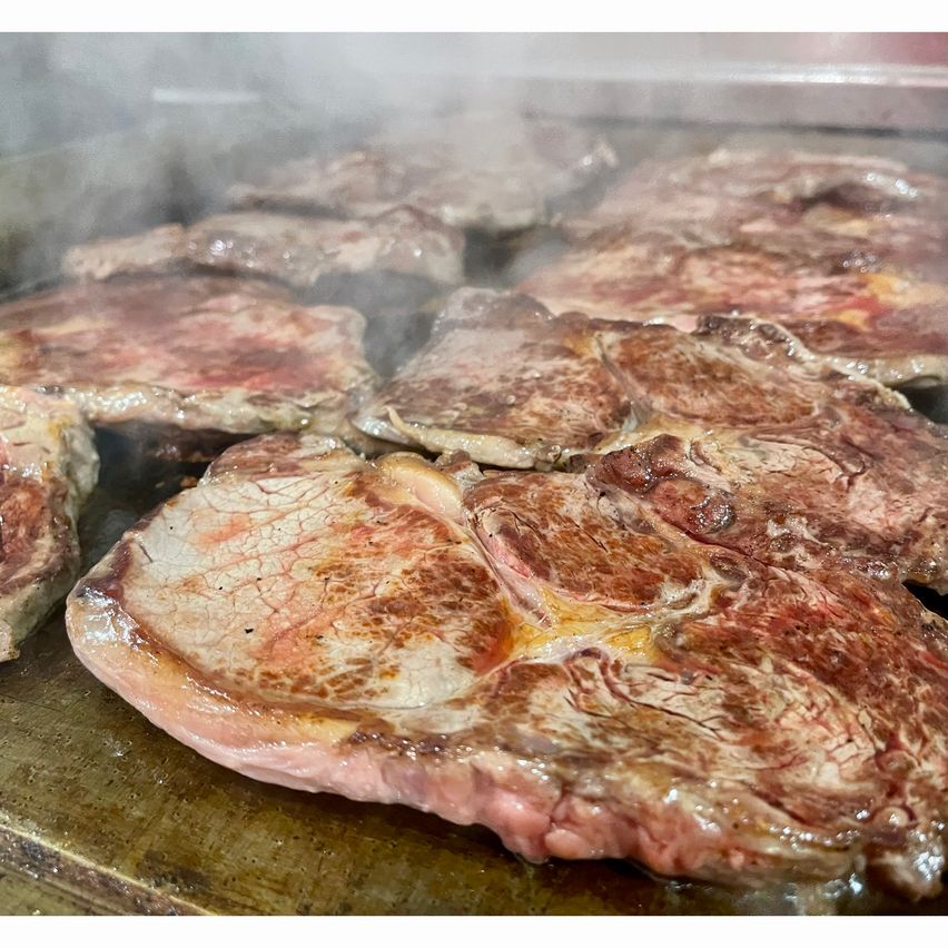 Mehrere Steaks werden auf einem Grill mit aufsteigendem Dampf gekocht. Die Steaks sind braun und haben eine marmorierte Textur, was auf eine gute Fleischqualität hinweist.
