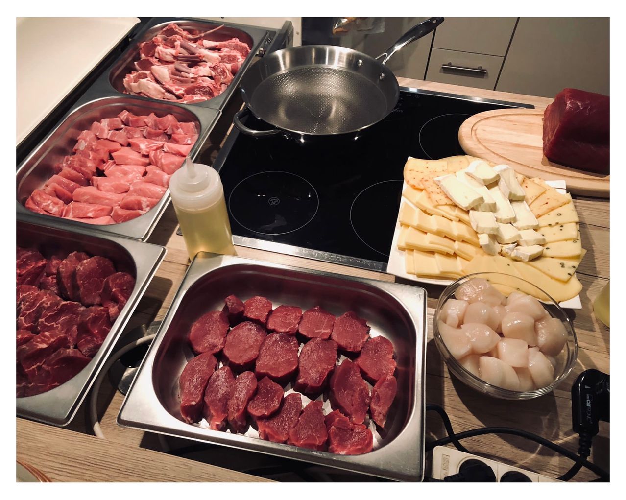Raw meat in trays, cheese, scallops, and a pan on a stove. Ingredients ready for cooking.