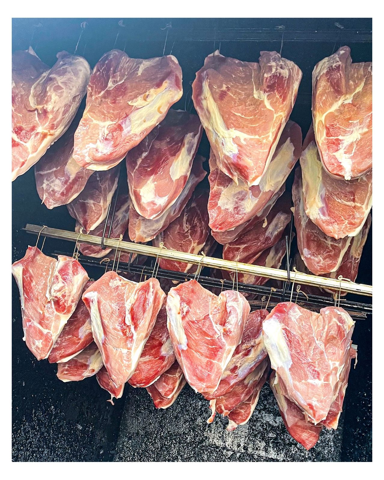 Raw pork chops are hanging on hooks inside a smoker, arranged in neat rows. The cuts exhibit a marbling of fat and lean meat, indicating quality.