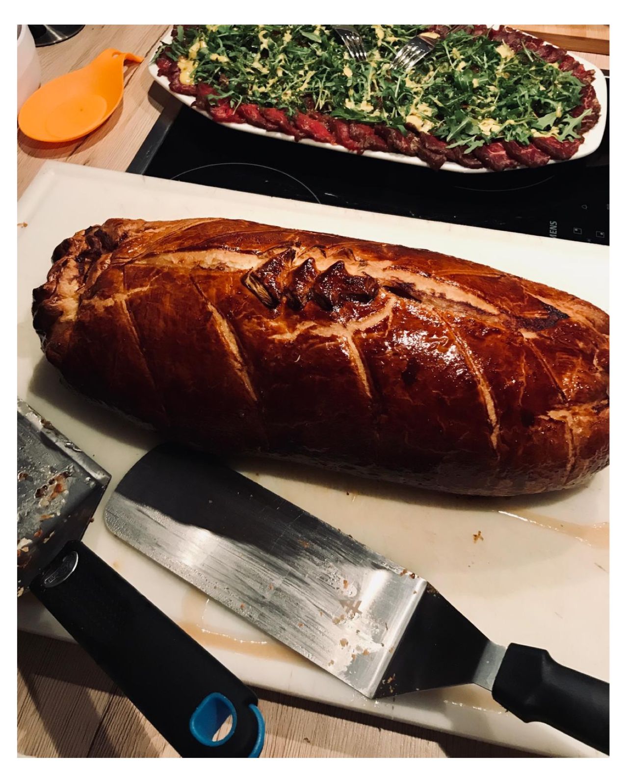 A large loaf of bread with cuts on its crust, placed on a white cutting board with two knives. Behind it, a steak with greens is on a plate over a stove.