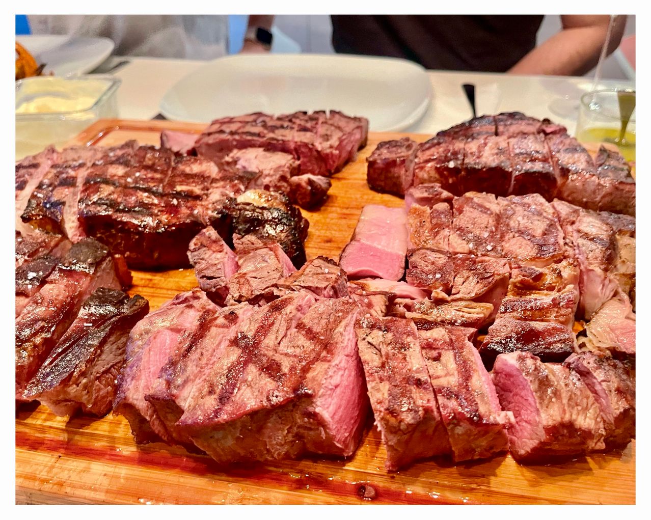 Auf einem Holzschneidebrett sind verschiedene gegrillte Fleischsorten zu sehen, darunter Steak und Rippchen, einige geschnitten und andere ganz. In der Nähe befinden sich Teller und Besteck.