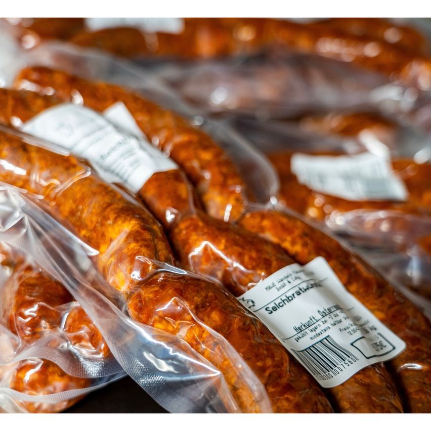 Nahaufnahme mehrerer Wurstpackungen in durchsichtigem Plastik, jede mit einem weißen Etikett, ordentlich in einer Reihe auf einer dunklen Oberfläche gestapelt.