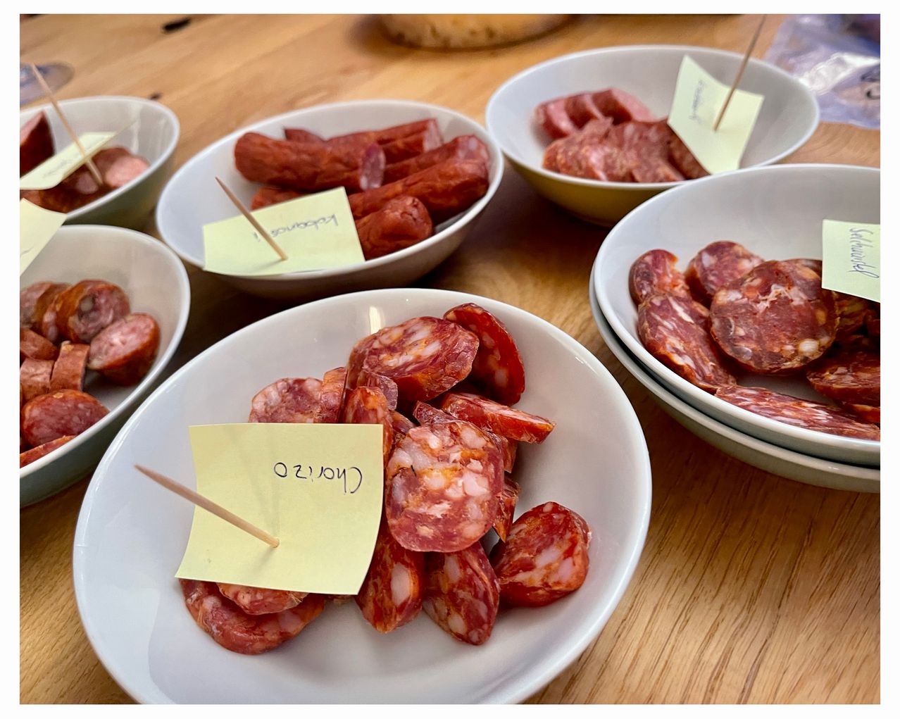 Mehrere weiße Schüsseln auf einem Holztisch enthalten verschiedene Arten von Würsten, jede mit gelben Etiketten. Die Schüsseln haben Zahnstocher, und die vorderste Schüssel ist prominent mit Chorizo beschriftet.