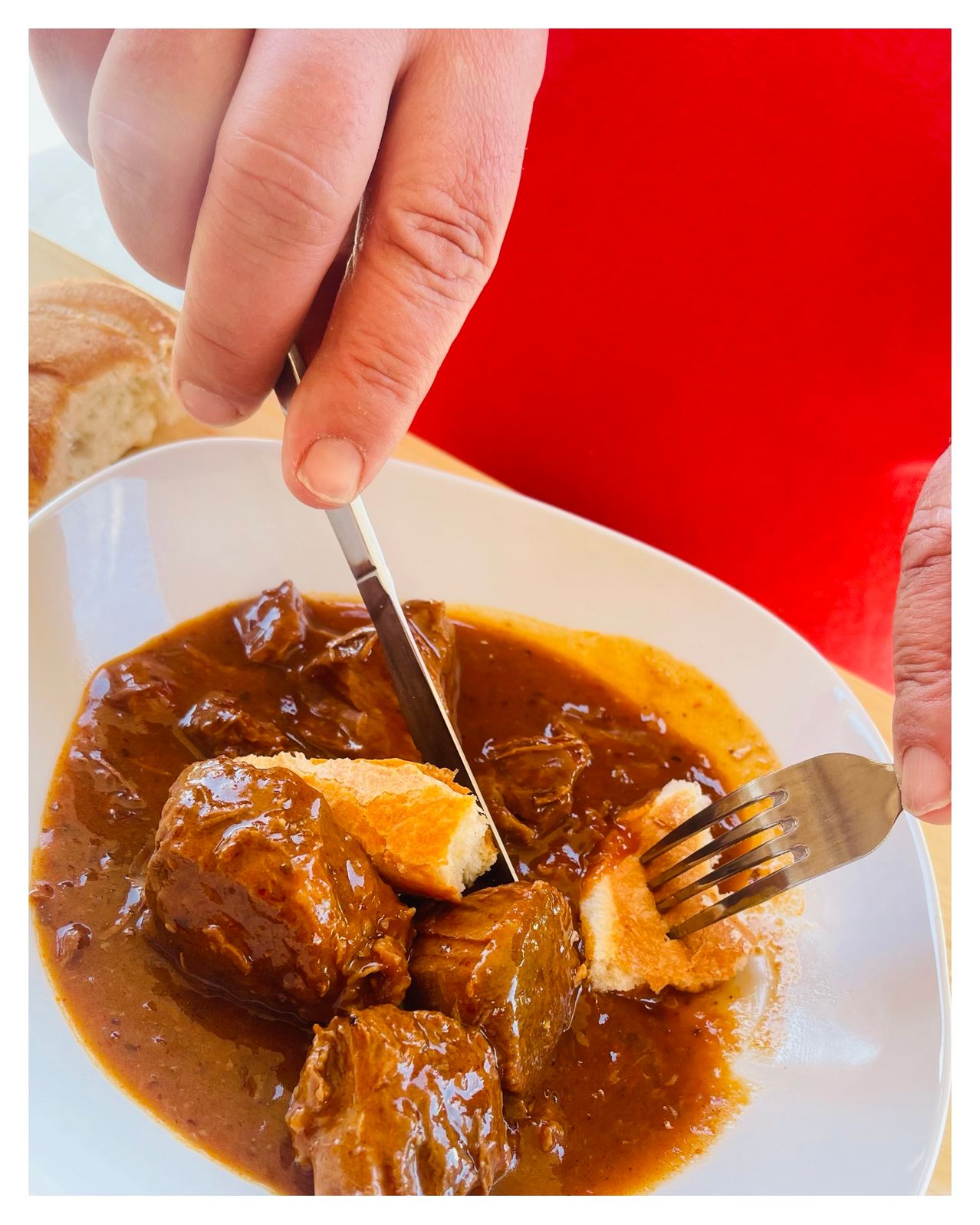 Eine Person schneidet ein Stück Fleisch aus einer Schüssel mit Eintopf mit Messer und Gabel, mit Brot auf der Seite.