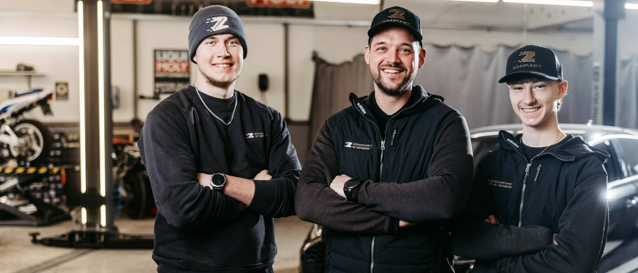 Zwei Männer in schwarzen Jacken und Mützen stehen in einer Garage, lächeln und haben die Arme verschränkt. Hinter ihnen sind ein Auto und verschiedene Garagenwerkzeuge zu sehen.