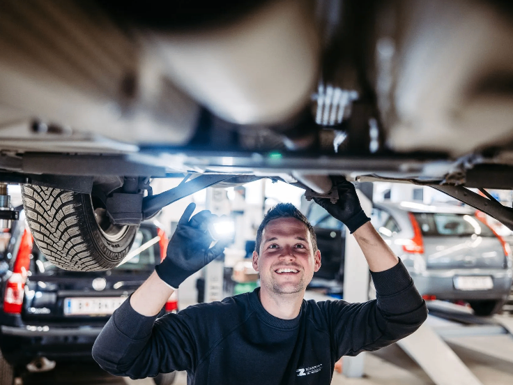 Ein Mann arbeitet an einem Auto unter einer Hebebühne, lächelt und hält eine Taschenlampe.