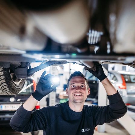 Ein Mann arbeitet an einem Auto unter einer Hebebühne, lächelt und hält eine Taschenlampe.