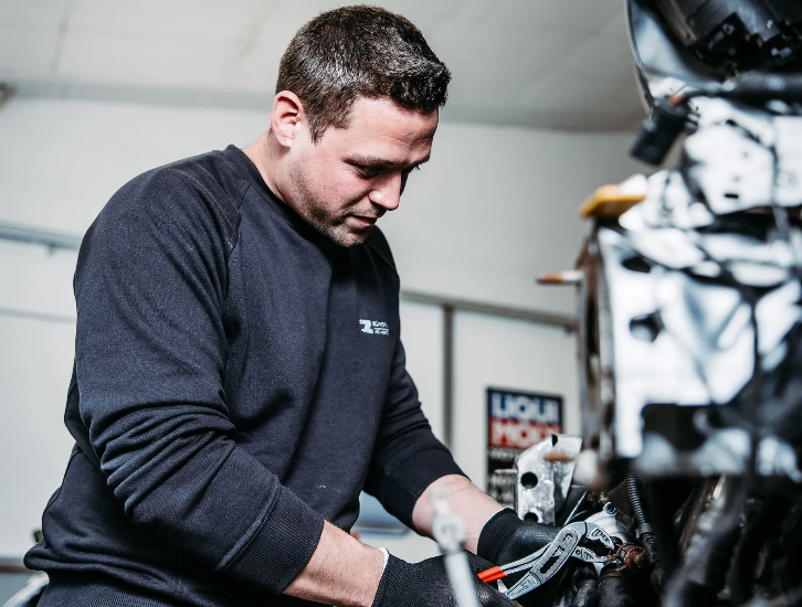 Ein Mann in einem blauen Shirt arbeitet an einem Motorradmotor. Er trägt Handschuhe und benutzt Werkzeuge.