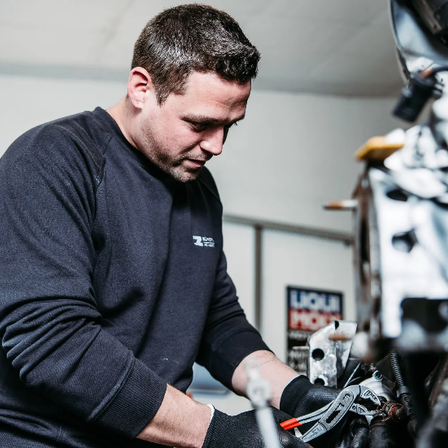 Ein Mann in einem blauen Shirt arbeitet an einem Motorradmotor. Er trägt Handschuhe und benutzt Werkzeuge.