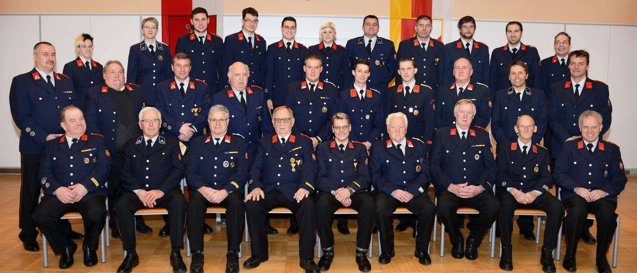 Gruppenfoto einer Feuerwehrmannschaft in Uniform, sitzend und stehend in einem Raum mit Fahnen im Hintergrund.