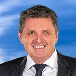 Ein Mann in formeller Kleidung lächelt in die Kamera, mit einem blauen Himmel im Hintergrund.
