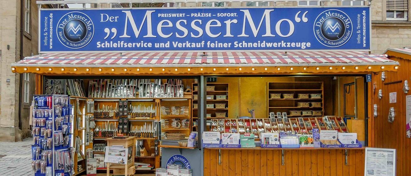 Ein Geschäft mit einem blauen Vordach namens 'Der MesserMo' verkauft feine Schneidwerkzeuge und bietet Schärfdienste an. Im Inneren zeigen Regale verschiedene Messer und Werkzeuge.