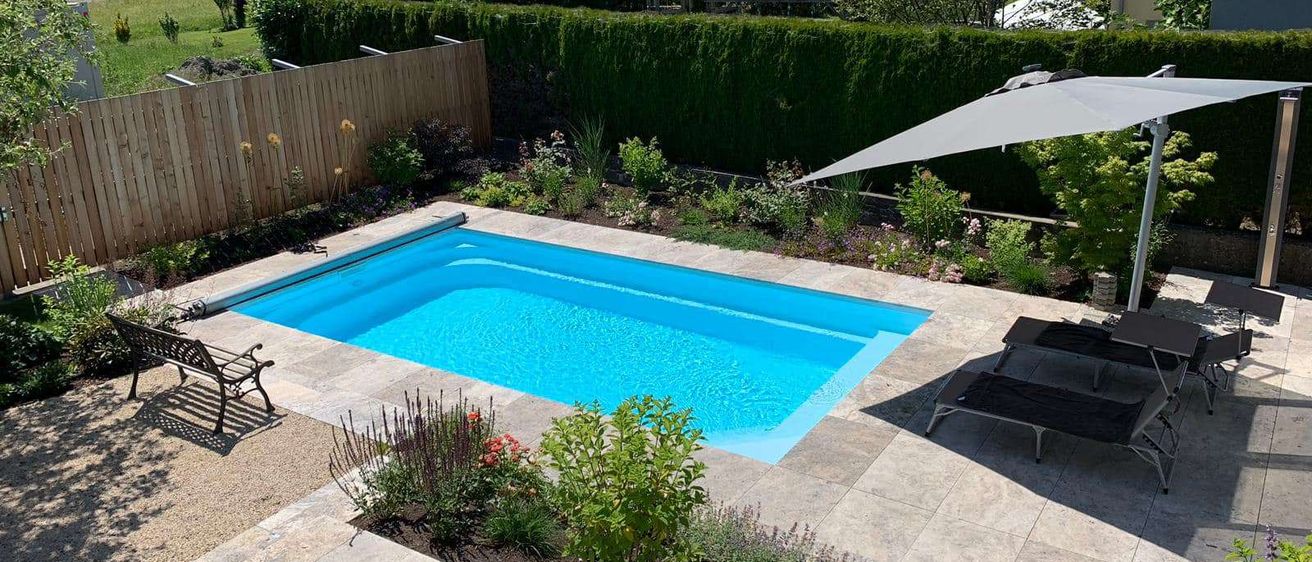Ein rechteckiger Swimmingpool befindet sich in der Mitte eines Hinterhofs, umgeben von Steinboden und Grünpflanzen. Links steht eine Holzbank und rechts ein Liegestuhl unter einem Schirm. Hinter dem Pool steht ein Holzzaun, und ein Haus ist in der Ferne.