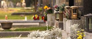 Ein Friedhof mit mehreren Grabsteinen und Gräbern. Auf einigen Gräbern stehen Blumen und brennende Laternen. Bäume und Gras sind im Hintergrund.