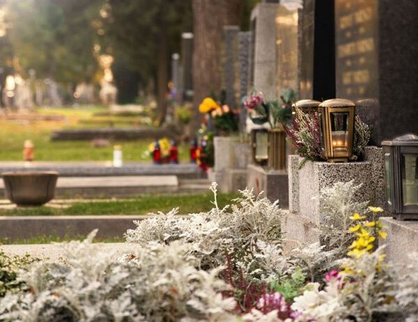 Ein Friedhof mit mehreren Grabsteinen und Gräbern. Auf einigen Gräbern stehen Blumen und brennende Laternen. Bäume und Gras sind im Hintergrund.