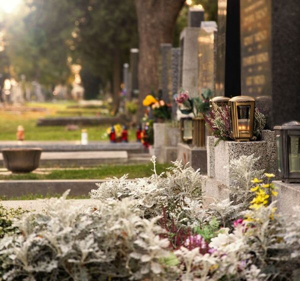 Ein Friedhof mit mehreren Grabsteinen und Gräbern. Auf einigen Gräbern stehen Blumen und brennende Laternen. Bäume und Gras sind im Hintergrund.