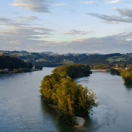 Ein ruhiger Blick auf einen Fluss mit einer kleinen Insel aus grünen Bäumen. Berge und sanfte Hügel säumen beide Seiten des Flusses unter einem bewölkten Himmel.