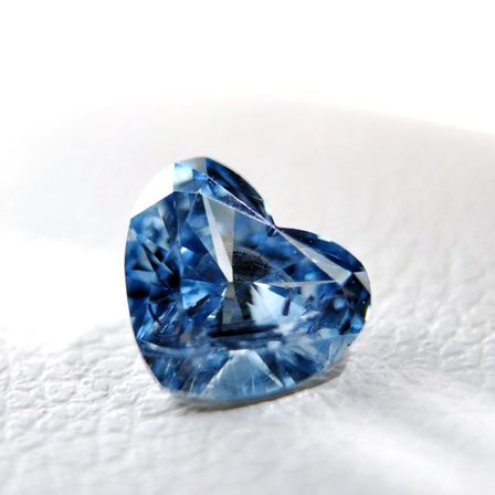 Ein Nahaufnahme eines blauen herzförmigen Edelsteins, möglicherweise ein Saphir, wird auf einer weißen Oberfläche gezeigt. Der Edelstein ist poliert und hat einen funkelnden Effekt.