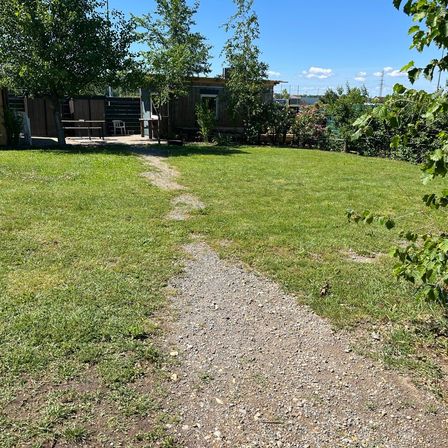 Ein Kiesweg führt durch einen Rasenbereich mit einem kleinen Holzhaus und einer Bank im Hintergrund. Bäume und Büsche umgeben den Bereich.
