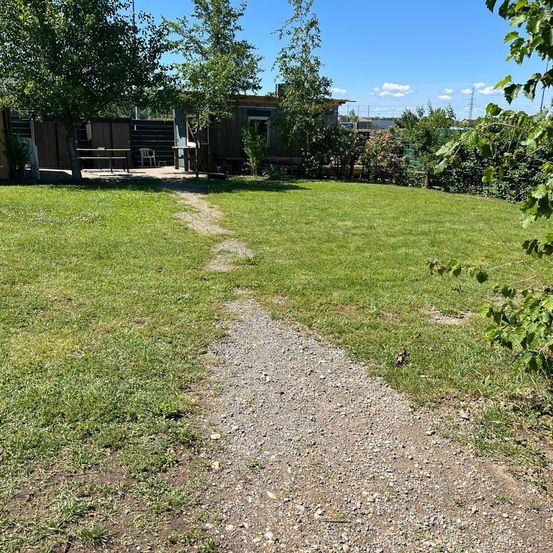 Ein Kiesweg führt durch einen Rasenbereich mit einem kleinen Holzhaus und einer Bank im Hintergrund. Bäume und Büsche umgeben den Bereich.