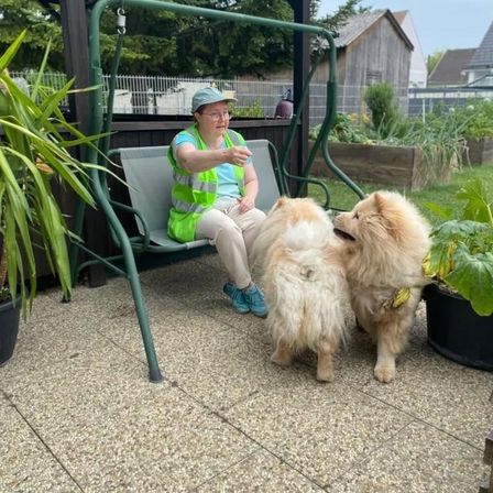 Eine Frau in einer grünen Weste sitzt auf einer Bank in einem Garten, umgeben von zwei Hunden, von denen einer eine Banane im Maul hat.