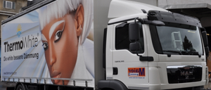 Ein weißer Lkw mit einer großen Werbung für ThermoWhite an der Seite, die eine Frau mit weißen Haaren zeigt. Der Lkw ist vor einem Gebäude geparkt.
