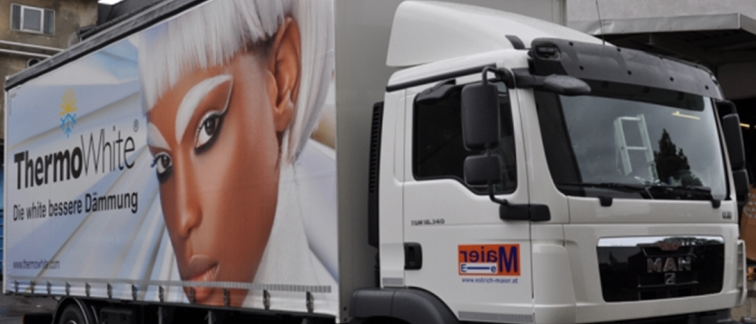 Ein weißer Lkw mit einer großen Werbung für ThermoWhite an der Seite, die eine Frau mit weißen Haaren zeigt. Der Lkw ist vor einem Gebäude geparkt.