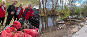 Vier Männer in roten Jacken posieren neben einem Auto mit Müllsäcken. Ein Fluss ist mit Trümmern am Ufer zu sehen.