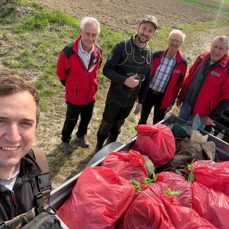Fünf Personen stehen in einem Feld mit mehreren roten Tüten in einem Truck. Ein Mann lächelt, während ein anderer zeigt. Die anderen tragen rote Jacken.