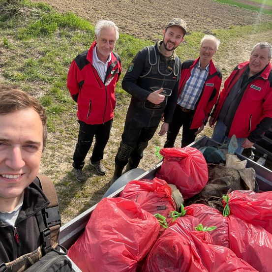 Fünf Personen stehen in einem Feld mit mehreren roten Tüten in einem Truck. Ein Mann lächelt, während ein anderer zeigt. Die anderen tragen rote Jacken.