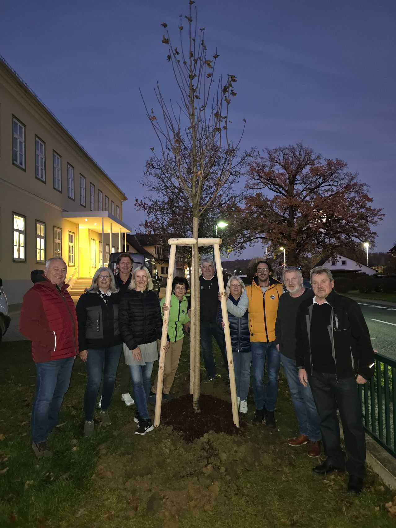 Eine Gruppe von Menschen steht um einen frisch gepflanzten Baum in der Dämmerung und lächelt und posiert für ein Foto vor einem Gebäude.