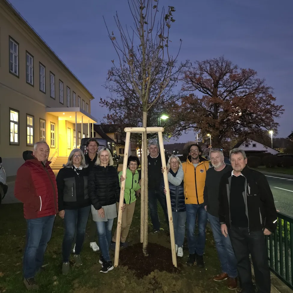 Eine Gruppe von Menschen steht um einen frisch gepflanzten Baum in der Dämmerung und lächelt und posiert für ein Foto vor einem Gebäude.