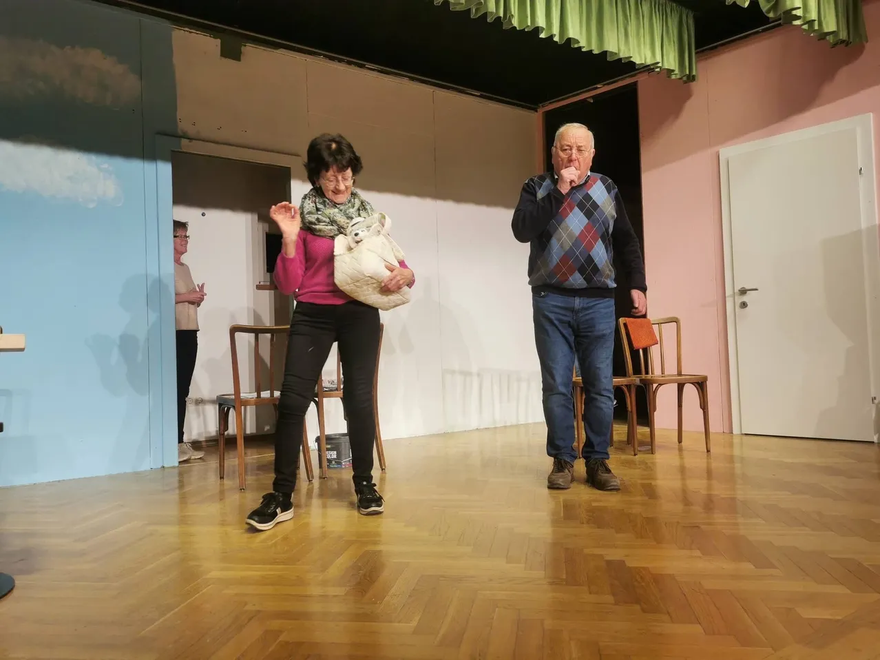 Eine Frau in einem rosa Pullover und schwarzen Hosen steht auf der Bühne mit einem Mann in einem karierten Pullover und Jeans. Sie hält einen Kuscheltier. Stühle und ein Eimer sind in der Nähe.