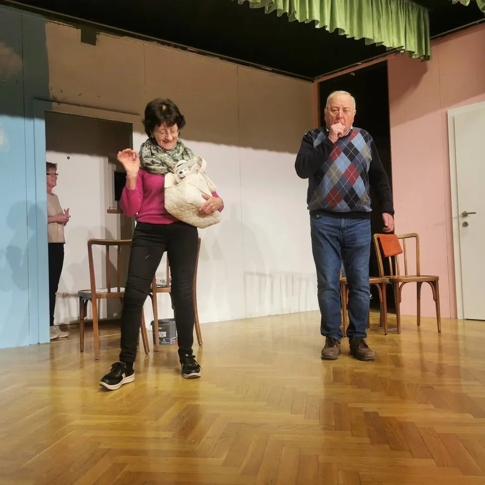 Eine Frau in einem rosa Pullover und schwarzen Hosen steht auf der Bühne mit einem Mann in einem karierten Pullover und Jeans. Sie hält einen Kuscheltier. Stühle und ein Eimer sind in der Nähe.