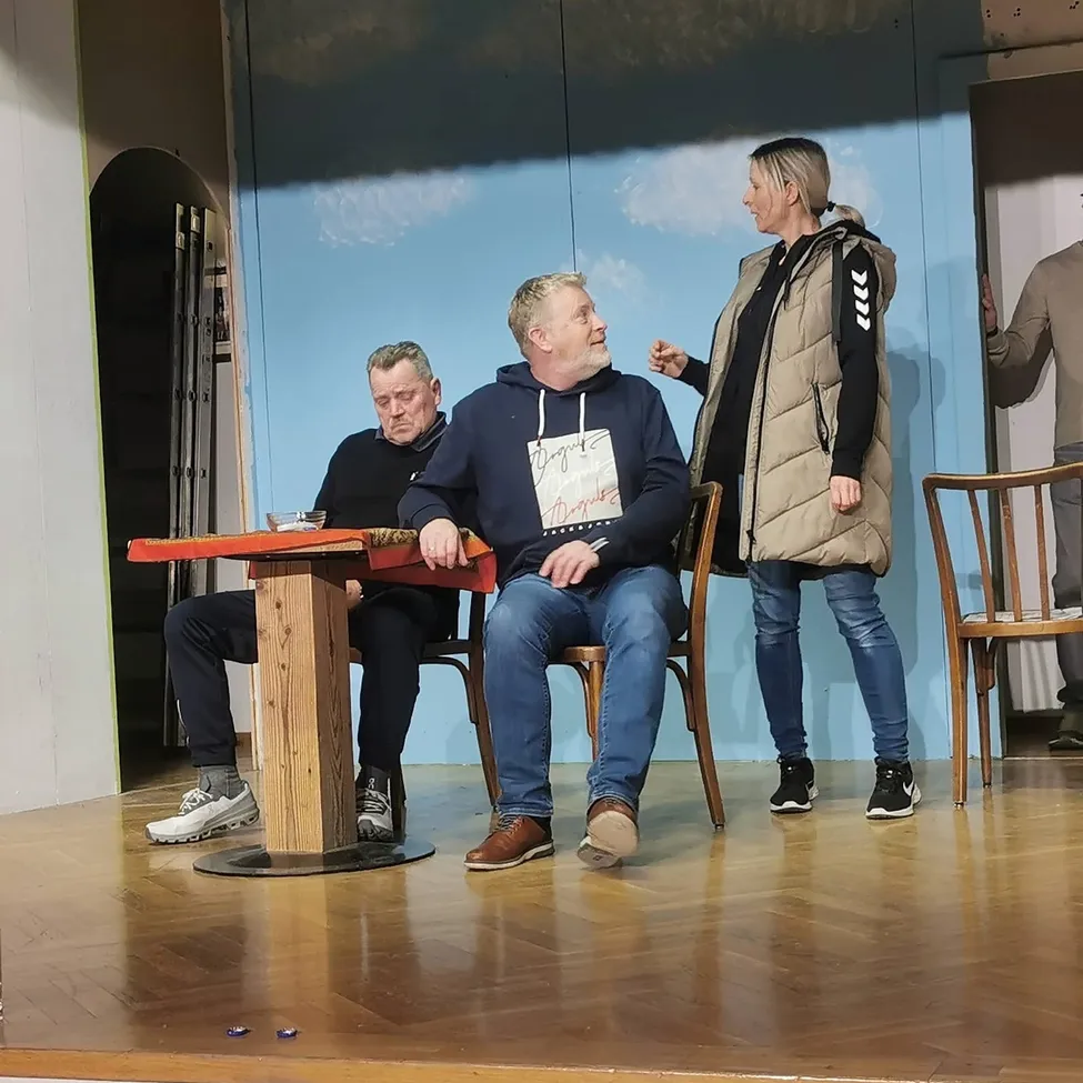 Drei Personen sind auf einer Bühne, auf Stühlen um einen Tisch herum sitzend. Ein Mann trägt eine Kapuzenjacke mit Text, ein anderer eine Jacke und Turnschuhe, und eine Frau in einer Jacke steht neben ihnen. Ein Wandbild ist hinter ihnen.