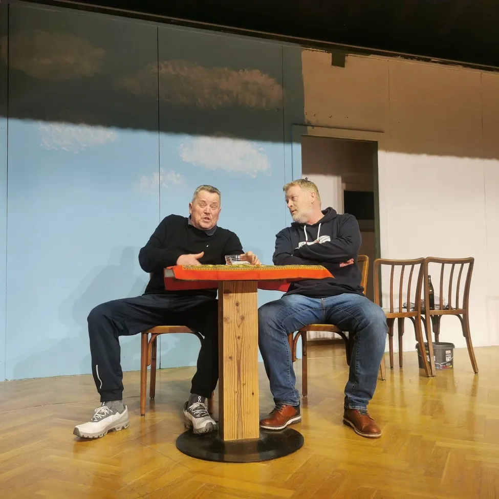 Zwei Männer sitzen sich gegenüber an einem Tisch in einem Raum mit einer blauen Wand. Ein Mann hat ein Glas, und beide haben die Arme verschränkt. Hinter ihnen befindet sich eine Tür, die zu einem anderen Raum führt.