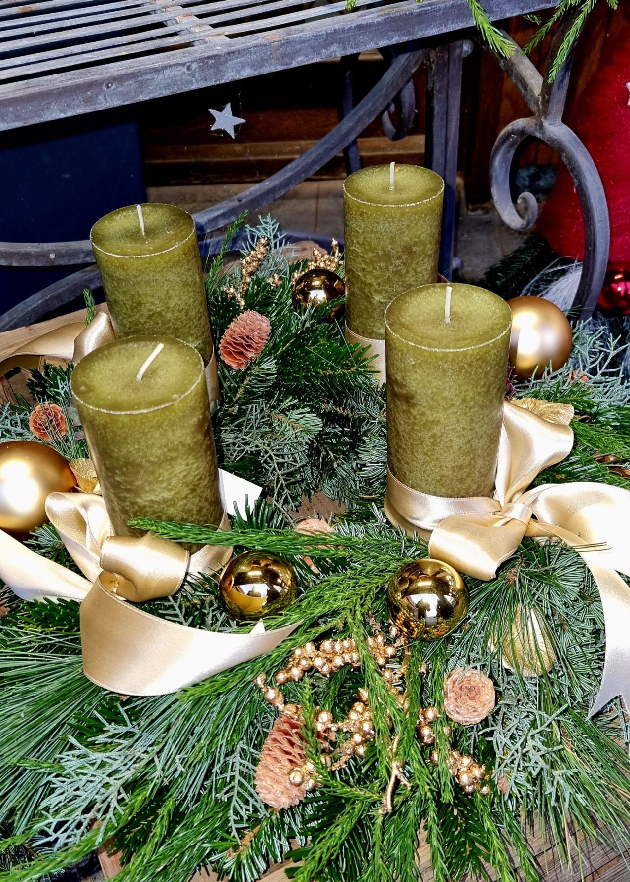 Ein Kranz aus grünen Kerzen, goldenen Ornamenten und Tannenzweigen liegt auf einer Holzoberfläche, geschmückt mit goldenen Bändern.