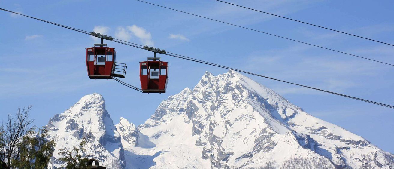 Zwei rote Seilbahnen hängen vor einem verschneiten Berg von einem Kabel ab. Das Kabel wird von zwei Türmen gestützt. Der Himmel ist klar mit einigen Wolken.