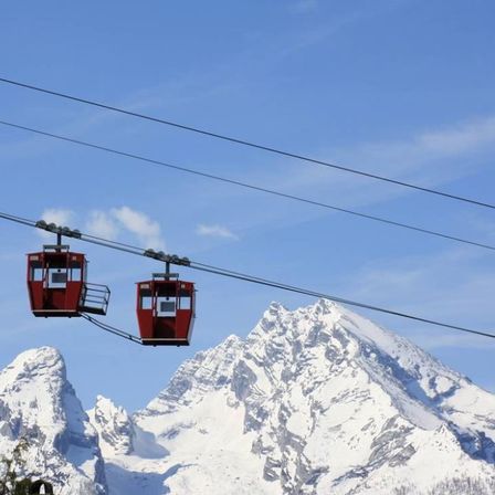 Zwei rote Gondelkabinen hängen von Seilen vor einem verschneiten Bergpanorama. Der Himmel ist klar mit einigen Wolken.