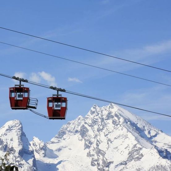 Zwei rote Gondelkabinen hängen von Seilen vor einem verschneiten Bergpanorama. Der Himmel ist klar mit einigen Wolken.