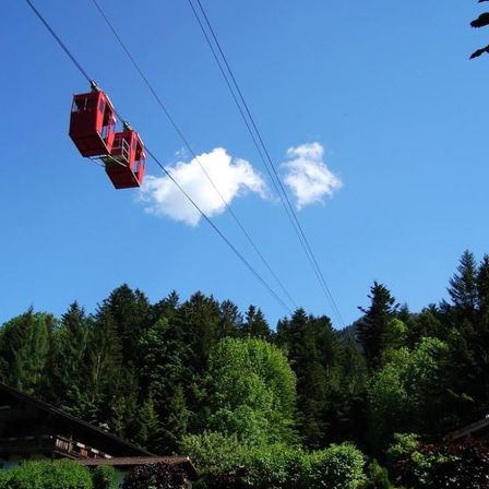 Eine rote Seilbahn fährt durch die Luft vor dem Hintergrund grüner Bäume und eines blauen Himmels mit einer Wolke.