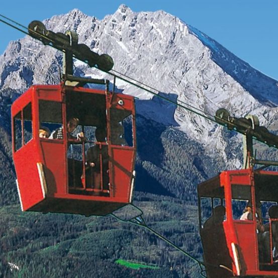 Zwei rote Seilbahnkabinen fahren einen Berg mit schneebedeckten Gipfeln hinauf. Passagiere im Inneren, einige schauen auf die Aussicht.