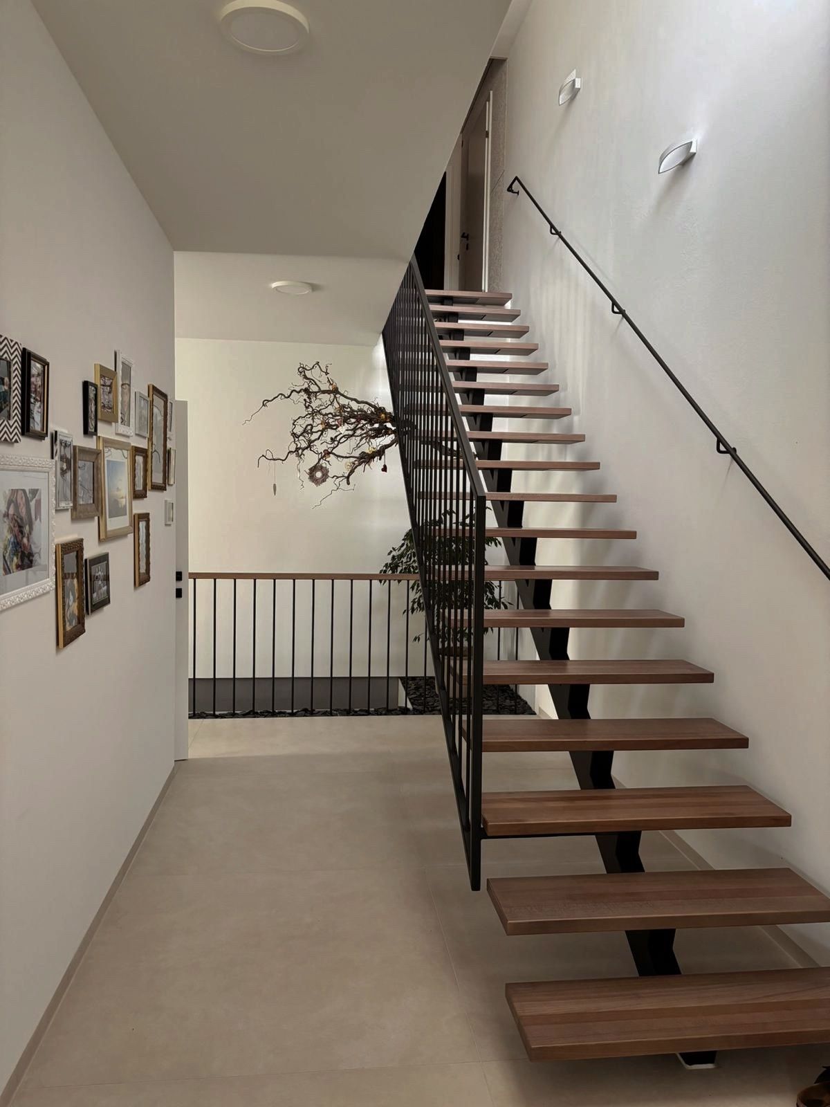 Ein Flur zeigt eine Holztreppe mit schwarzen Geländern und einer mit Bilderrahmen geschmückten Wand. Unter der Treppe steht ein Topf mit einer Pflanze.