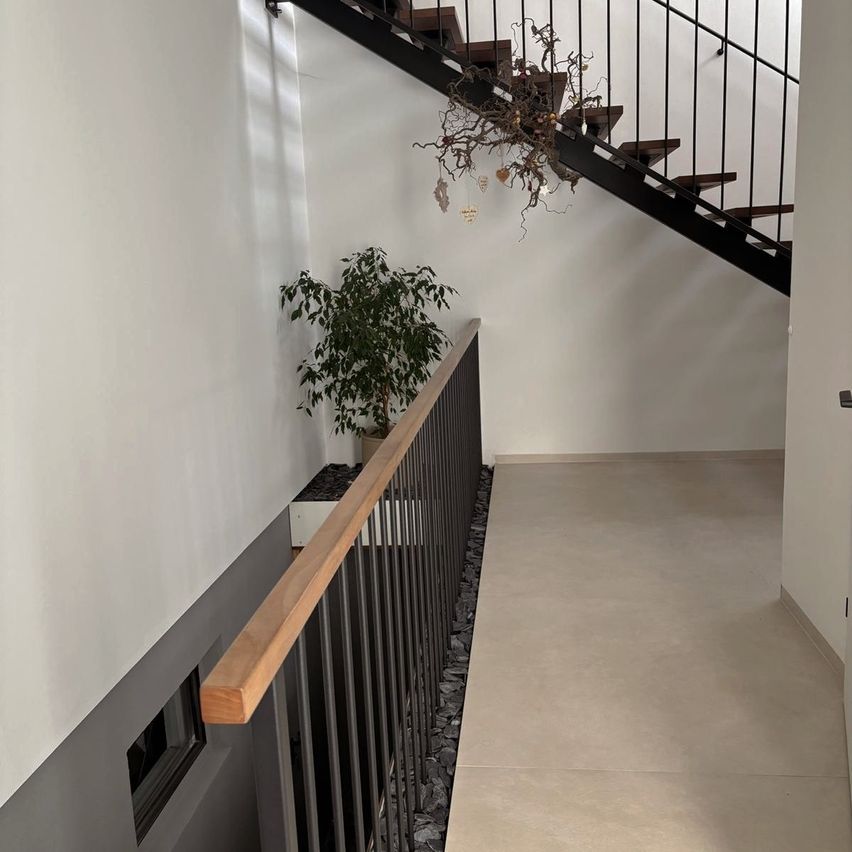 Ein moderner Innenflur mit einem Holzgeländer. Eine Pflanze ist neben einer Treppe platziert. Über der Treppe hängt ein dekoratives Element.