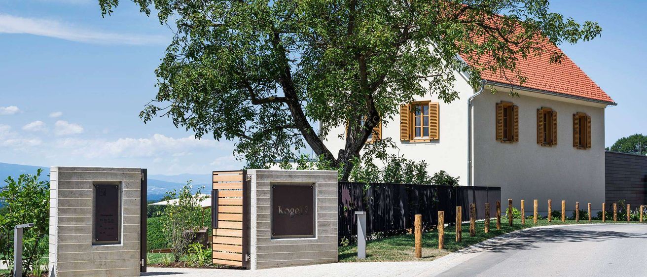 Ein Haus mit rotem Dach und gelben Fensterläden steht unter einem großen Baum an einem sonnigen Tag. Der Baum ist groß und hat grüne Blätter.