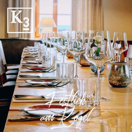 Ein langer Esstisch ist mit vielen Gläsern, Tellern und Besteck gedeckt. Jede Tischplatzierung beinhaltet Gabel, Messer und Löffel. Der Tisch ist mit einer Blume in einer Vase dekoriert.