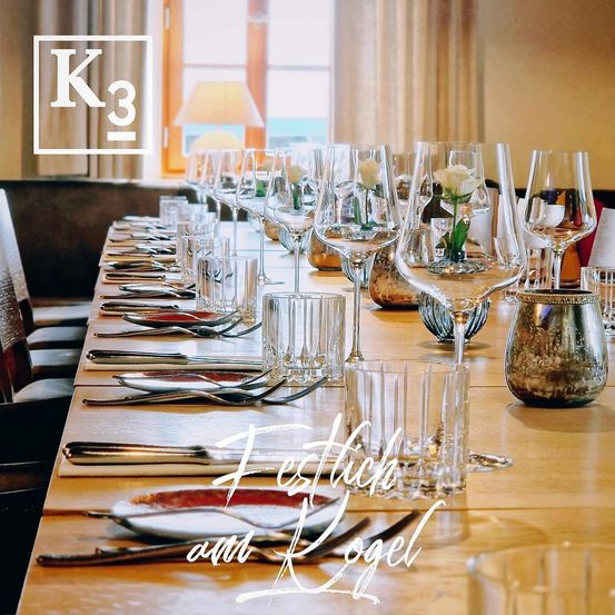 Ein langer Esstisch ist mit vielen Gläsern, Tellern und Besteck gedeckt. Jede Tischplatzierung beinhaltet Gabel, Messer und Löffel. Der Tisch ist mit einer Blume in einer Vase dekoriert.