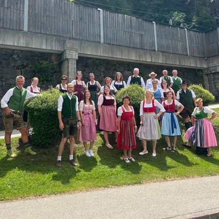 Eine Gruppe von Menschen in traditioneller deutscher Kleidung, einschließlich Lederhosen und Dirndln, steht vor einer Betonbrücke mit Grünanlagen und Bergen im Hintergrund.